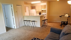A living room with a gray sofa, a patterned rug, and a kitchen area with a refrigerator, stove, and bar stools. - Condo near IMG Academy & Beaches (Bradenton)