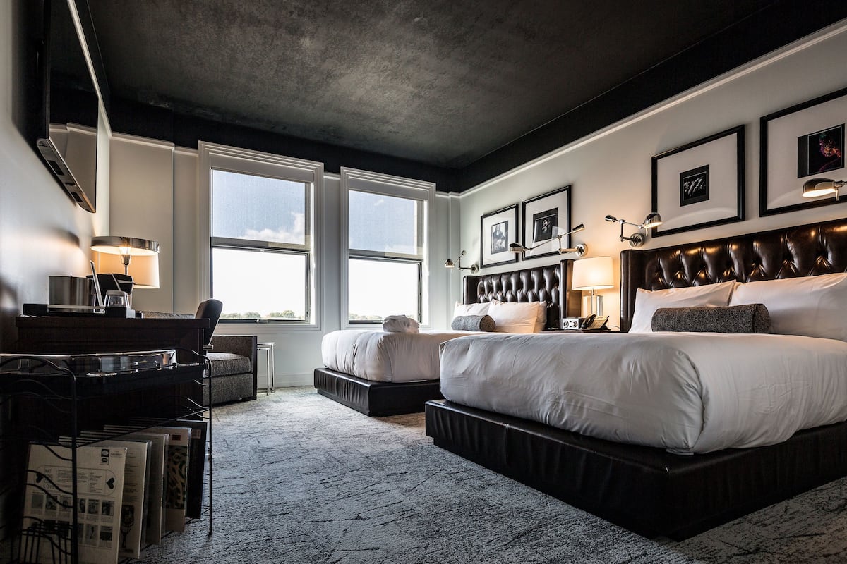 Habitación de hotel moderna con una cama grande, un escritorio y una silla. Hay dos ventanas que dejan entrar luz natural.