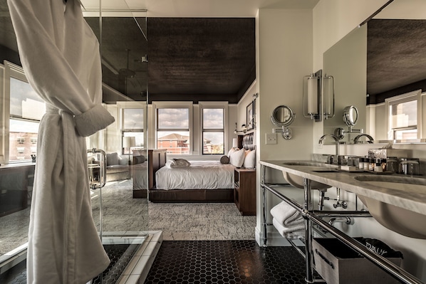 A modern hotel room with a large bed, a bathroom with a large mirror and sink, and a view of buildings outside. - Hotel Goodwin (Beloit)