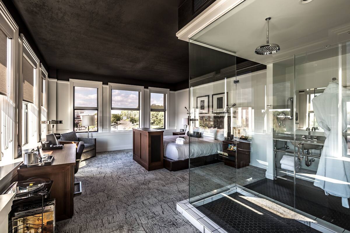 Un dormitorio moderno con una cama grande, un escritorio, cabina de ducha de vidrio y ventanas amplias que ofrecen vistas al exterior.