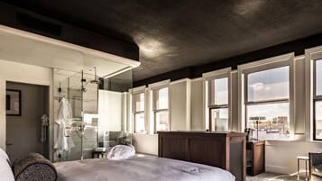 A modern bedroom with a large bed, a glass shower enclosure, and a view of the city through large windows.