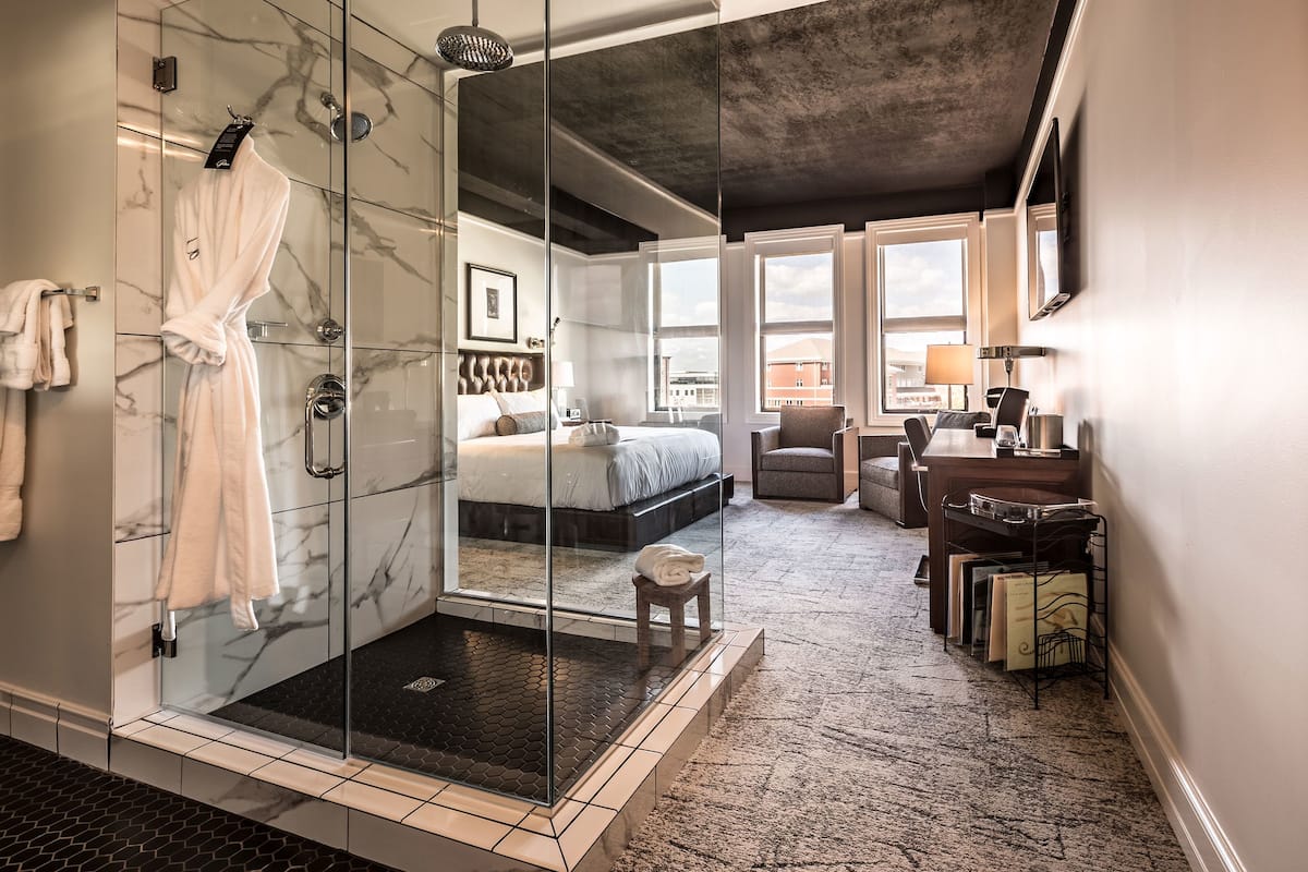 Habitación de hotel moderna con ducha de vidrio, cama grande, zona de estar y vistas a la ciudad.