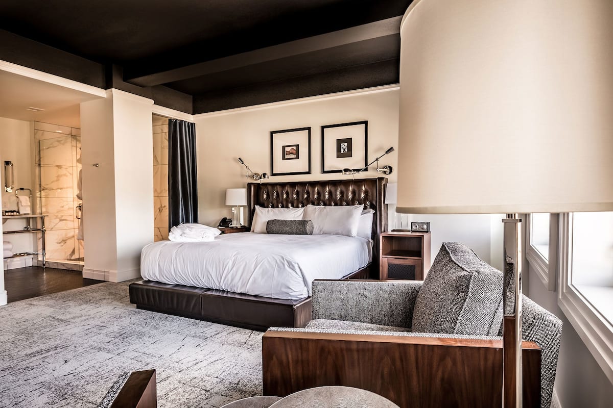 Una habitación de hotel con una cama grande, una zona de estar con sofá y sillón, una mesita de noche, una lámpara y un cuadro en la pared.