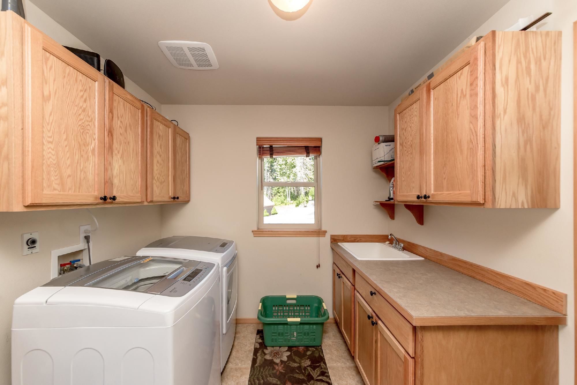 A compact kitchen with wooden cabinets, a washing machine, a sink, and a window with a view of greenery.