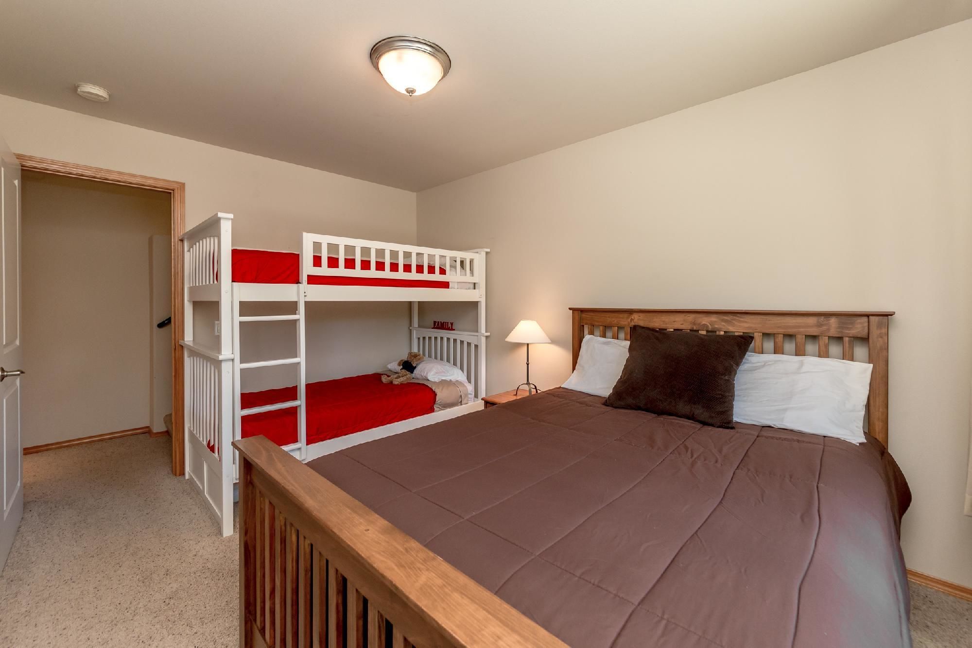 Une chambre avec un lit superposé, un lit simple, une table de chevet avec une lampe et une porte donnant sur une autre pièce.