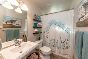 Un baño con un lavamanos blanco, un inodoro y una cortina de ducha con estampado de lluvia. Hay repisas con artículos de aseo personal y un cuadro de un granero en la pared.