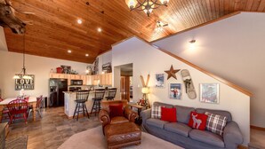 A spacious living area with a kitchen, a dining table with red chairs, a leather sofa, and a staircase leading to an upper level. - Wintergreen Chalet (Cle Elum)