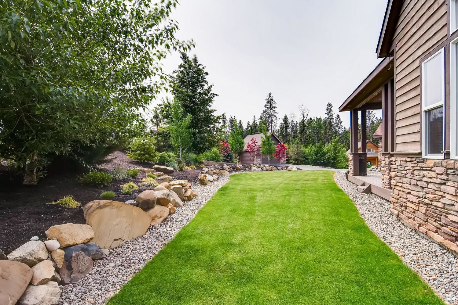 A well-maintained lawn with a stone border, a wooden house with a stone facade, and a forested backdrop.