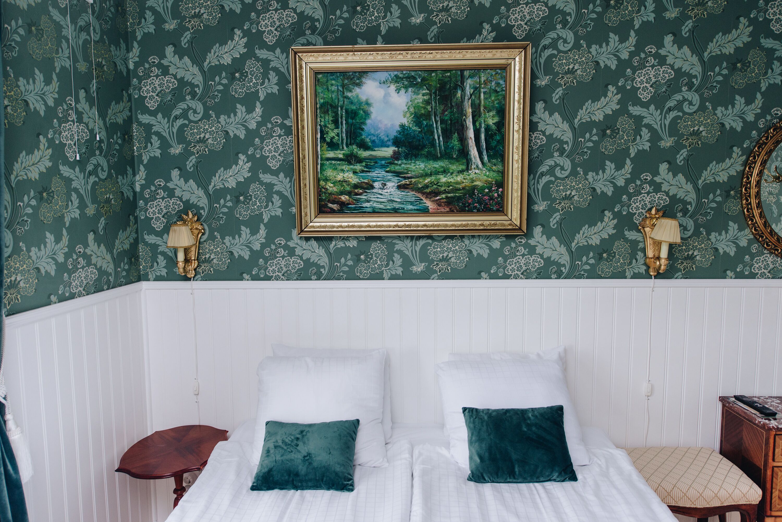 A bedroom with a bed, two pillows, a nightstand, a lamp, and a framed painting of a forest scene.