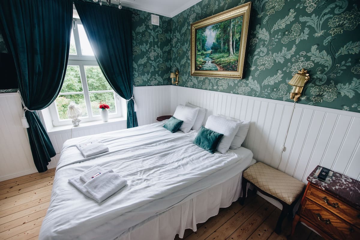 A neatly made bed with white linens and teal pillows, a bedside table with a lamp, and a window with curtains.
