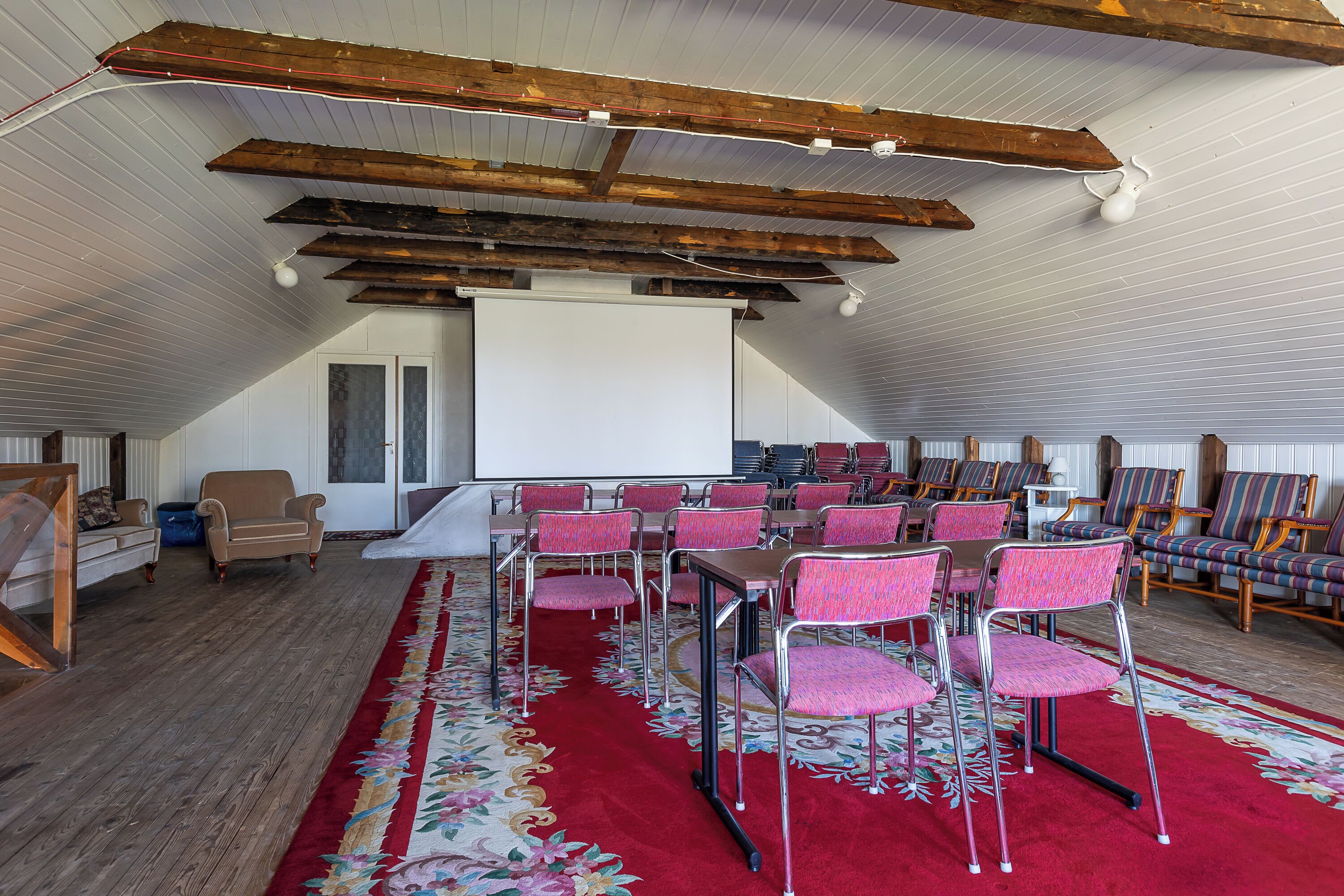 A spacious room with a high ceiling, wooden beams, a red carpet, and rows of pink chairs arranged for seating.
