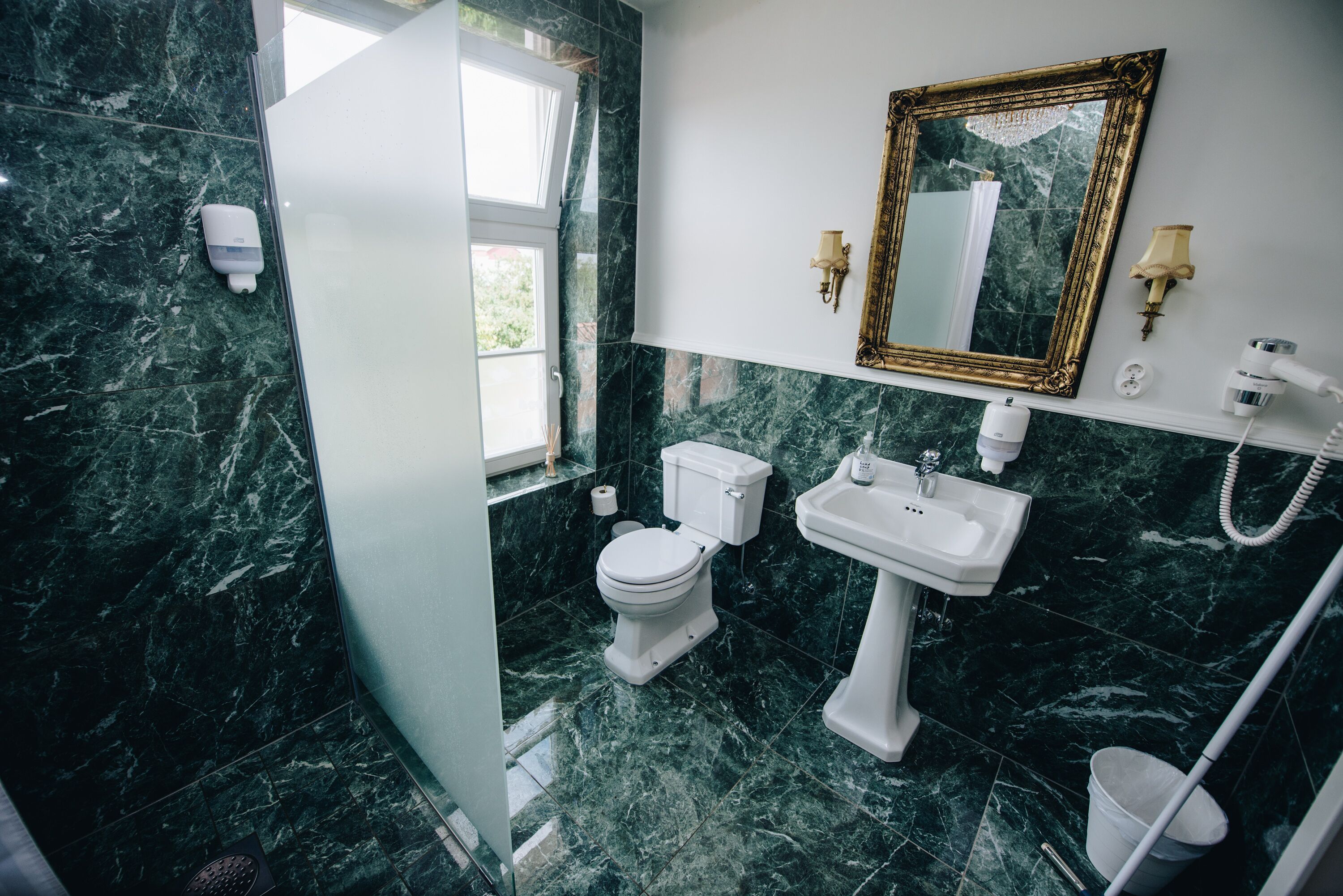 A bathroom with a toilet, sink, and mirror. There is a window providing natural light and a shower area.