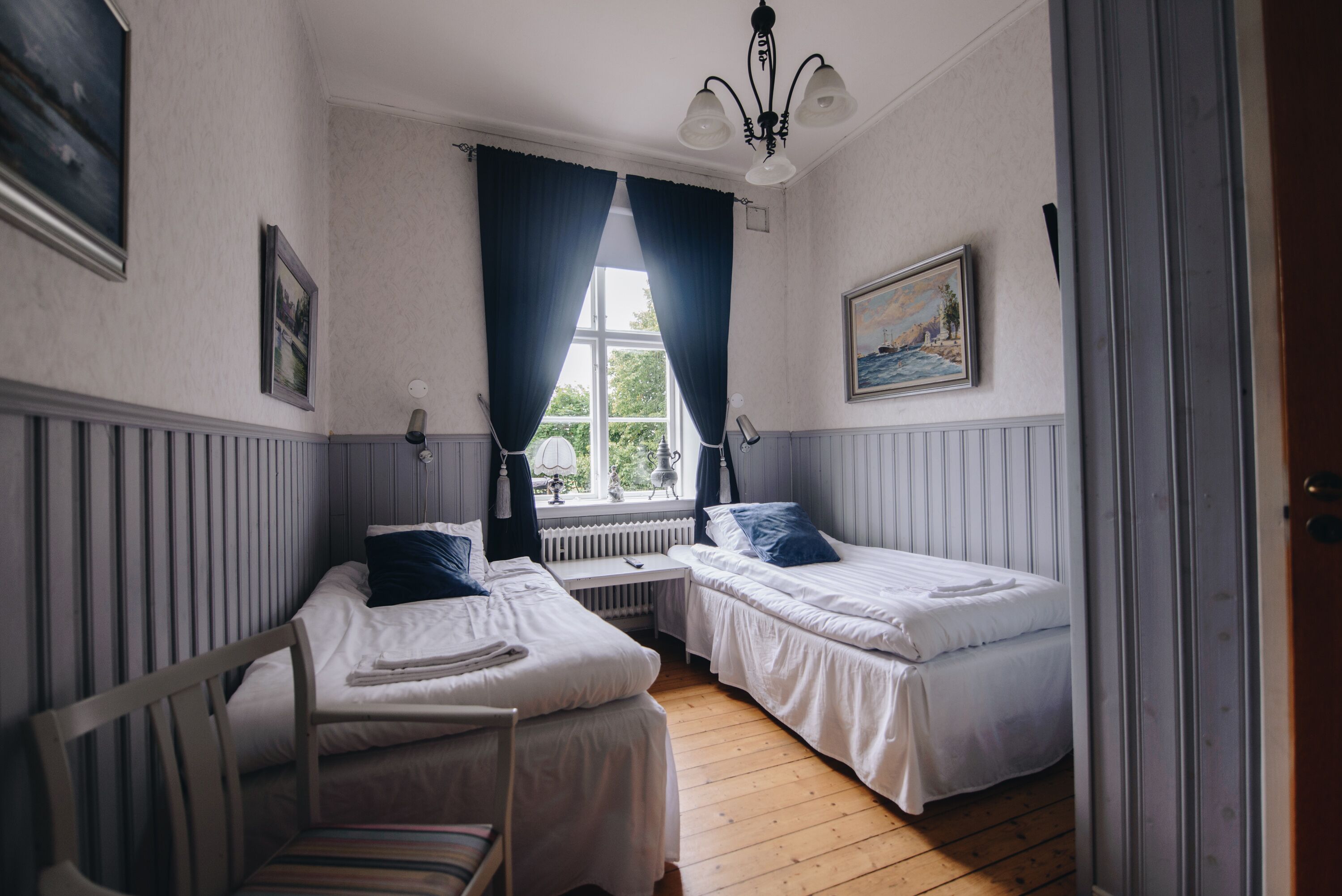 Una habitación con dos camas individuales, una silla, una mesa, un ventanal con cortinas, un cuadro en la pared y un radiador.