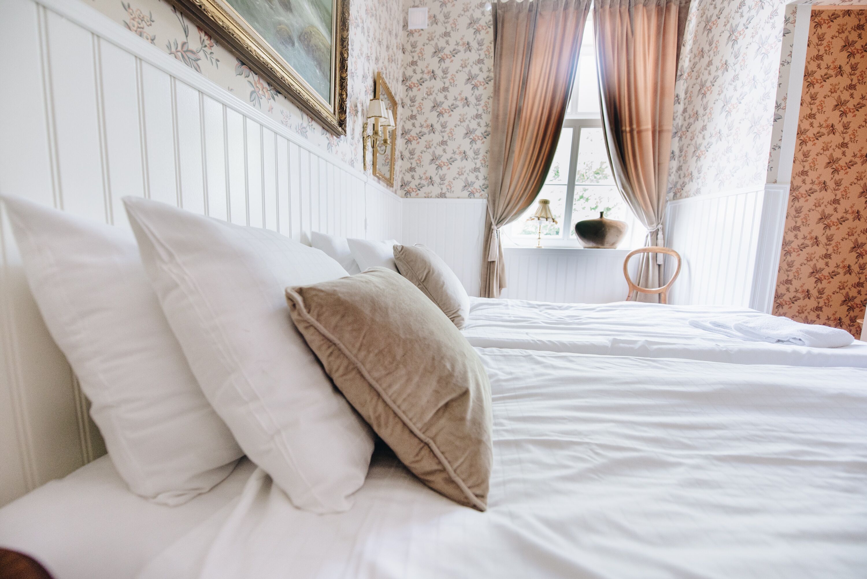 Una cama bien tendida con almohadas blancas y color beige, una silla de madera y una ventana con cortinas.