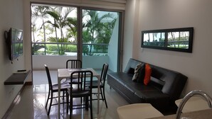 A modern living room with a dining area, large windows with a view of palm trees, a black leather sofa with cushions, and a flat-screen TV mounted on the wall. - Apartamento Mistral Cielo Mar (Cartagena)