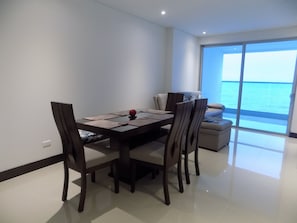 A dining area with a table and chairs, a sofa, and a view of the sea through a large window.