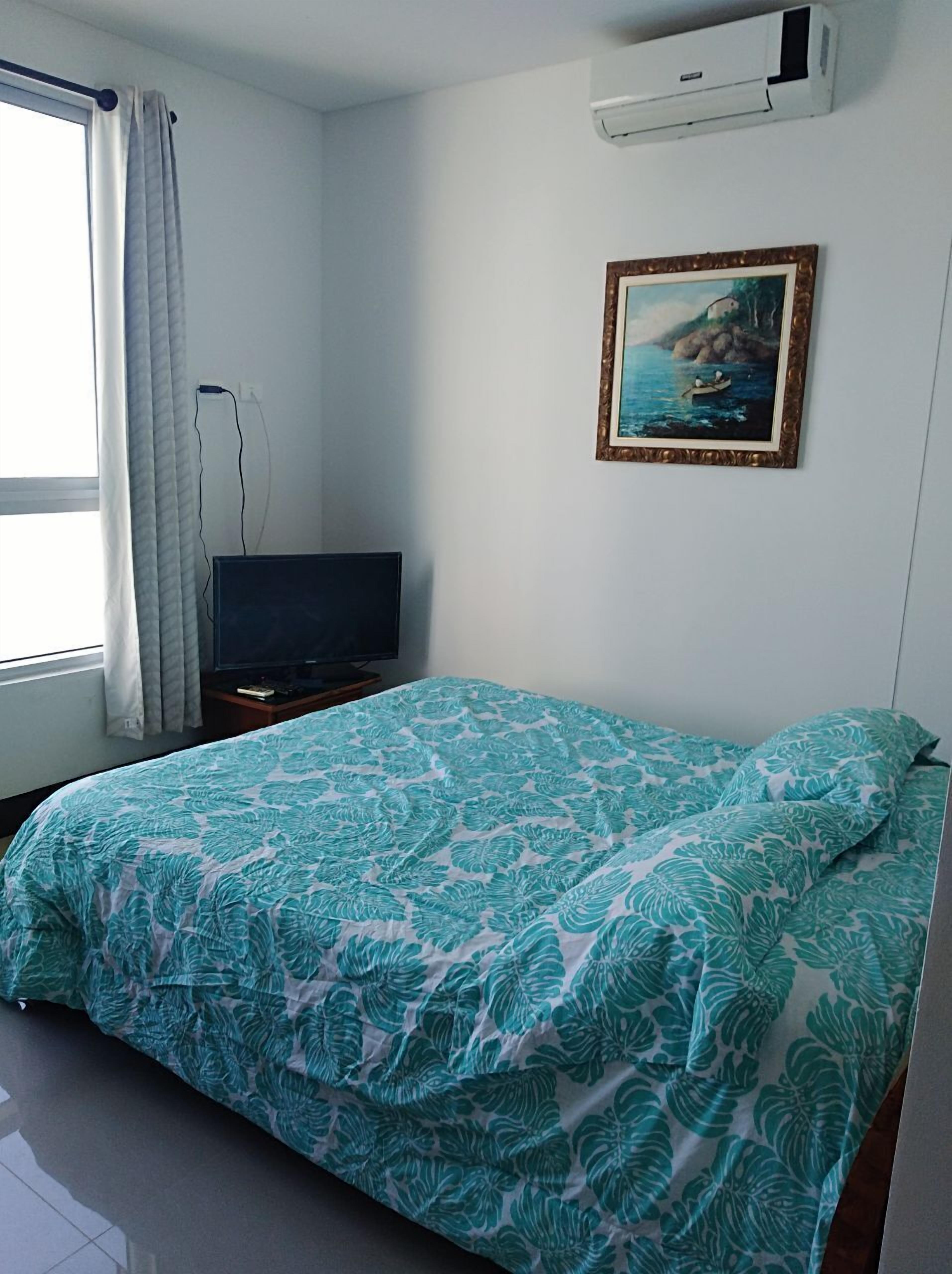 A bedroom with a bed, a television on a stand, a painting on the wall, and an air conditioner.
