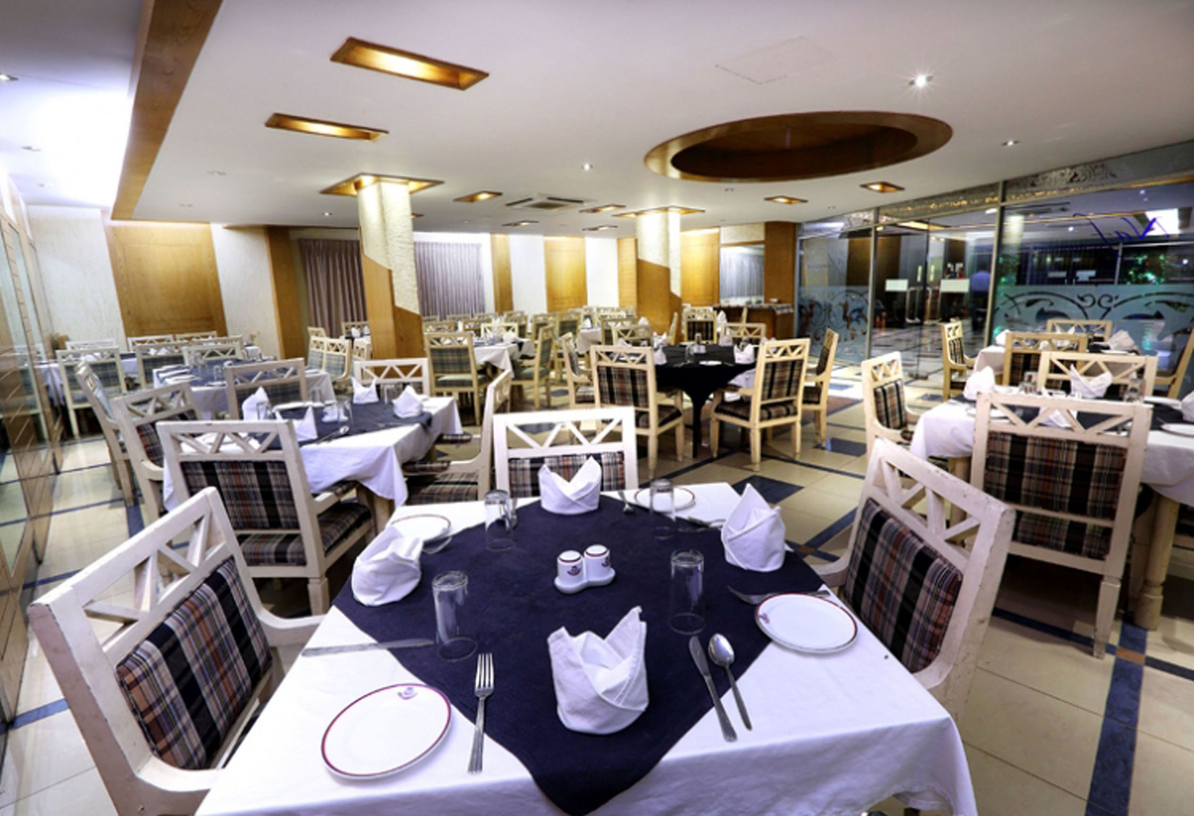 A dining area with round tables set for four, each with white tablecloths, napkins, cutlery, and glasses. The chairs have plaid upholstery.