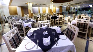 A dining area with round tables set for four, each with white tablecloths, napkins, cutlery, and glasses. The chairs have plaid upholstery. - Uni Resort (Cox's Bazar)