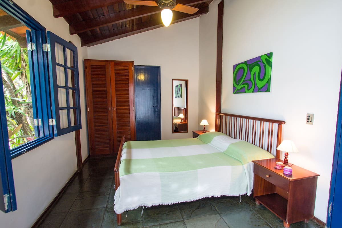 Quarto com cama, mesa de cabeceira, espelho fixado na parede e janela com persianas azuis.