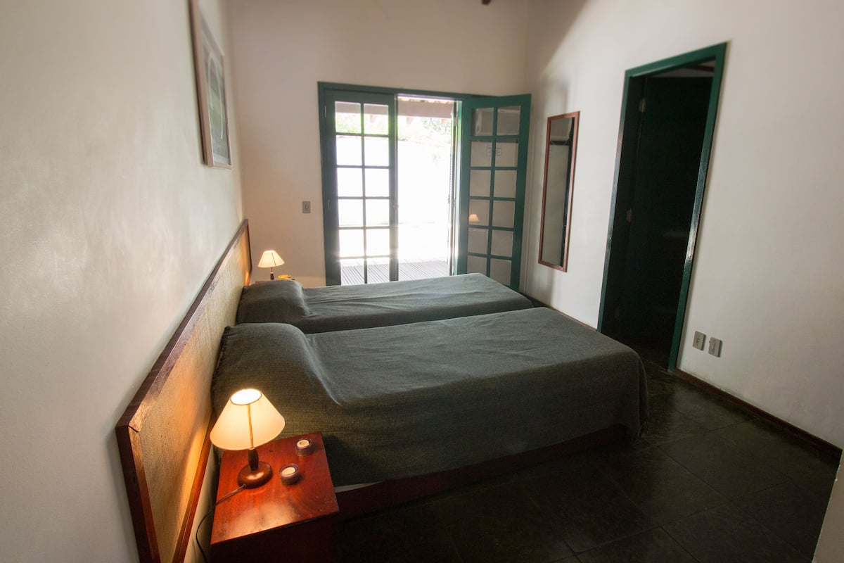Quarto com duas camas, uma mesa de cabeceira com abajur, uma porta e uma janela.