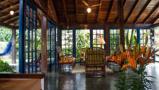 A spacious indoor area with a high ceiling, wooden beams, and large windows allowing natural light. There are several wicker chairs and a table, surrounded by potted plants and flowers. - Homestay Búzios (Búzios)