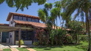 A two-story house with a tiled roof, a covered porch, and a well-maintained garden with palm trees and flowering plants. - Homestay Búzios (Búzios)