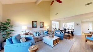 A spacious living room with a high ceiling, a dining area with a large window, and a kitchen visible in the background.