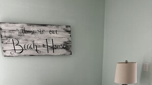 A room with a blue armchair, a wall-mounted sign reading "Welcome to our Beach House," a lamp on a side table, and a cushion with "BEACH" on it. - Quaint Condo Seagrove Beach right fof 30A, quick walk to two beach accessPo (Santa Rosa Beach)