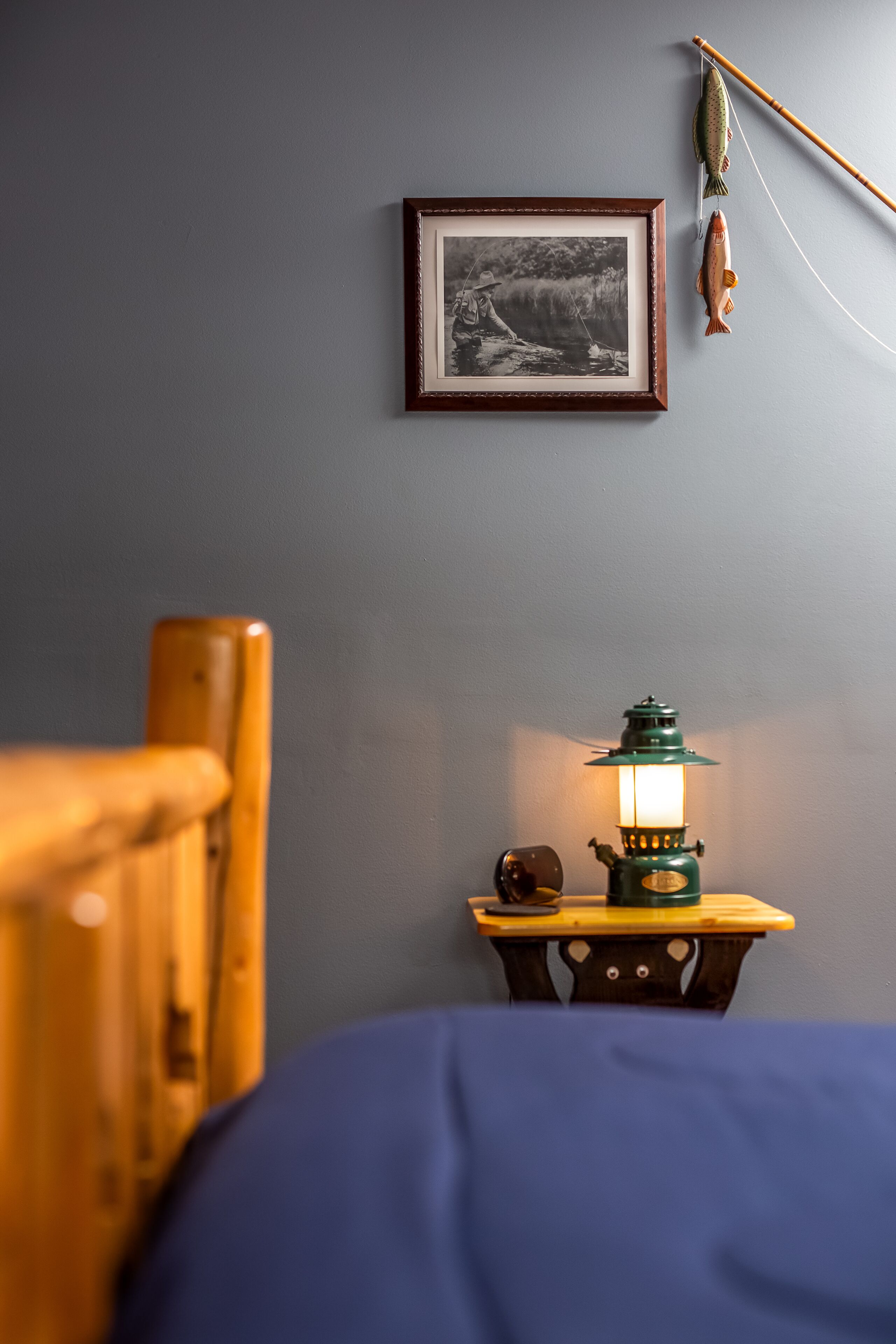 Une pièce avec une photographie encadrée accrochée au mur, une canne à pêche avec un poisson, une chaise en bois et une table sur laquelle se trouve une lanterne verte.