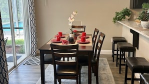 Een eetgedeelte met een houten tafel, stoelen en een kroonluchter. Er is een raam met jaloezieën en uitzicht op groen.