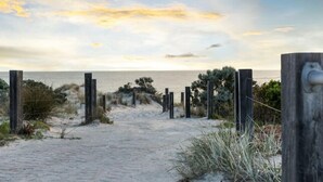 Un sentier de plage sablonneux, bordé de poteaux en bois et parsemé de quelques plantes, qui mène à la mer.