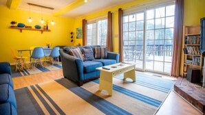 A bright living room with a blue sofa, a coffee table, and a dining area with chairs and a table. - Here Comes the Sun: 2 Kayaks, Wifi, Ping Pong & Pool table,Sandy Beach, Fenced in Yard, Pet Friendly (Huddleston)