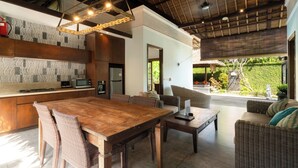 A spacious kitchen with a wooden table and wicker chairs, a dining area with a sofa and coffee table, and a view of a lush garden through large windows. - Kecapi Villa Seminyak (Seminyak)