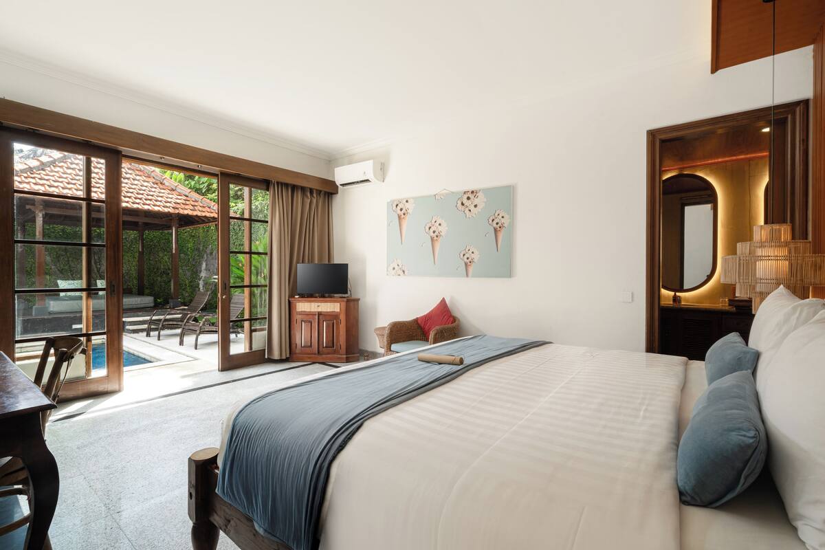 A hotel room with a large bed, a television, a painting, and a view of an outdoor pool area.