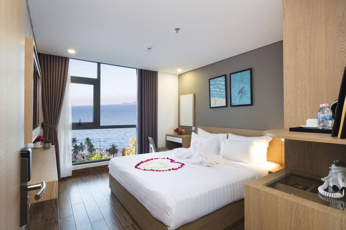 A hotel room with a large bed, a desk, a chair, a nightstand, and a view of the ocean.