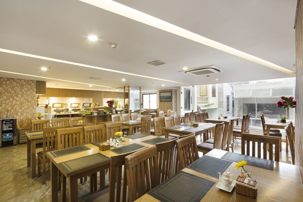 A spacious dining area with wooden tables and chairs, a kitchen in the background, and large windows allowing natural light. - Smile Hotel (Nha Trang)