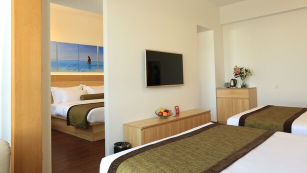 Une chambre d’hôtel avec deux lits, une télévision, un petit bureau avec un panier de fruits et un tableau représentant une scène de plage.