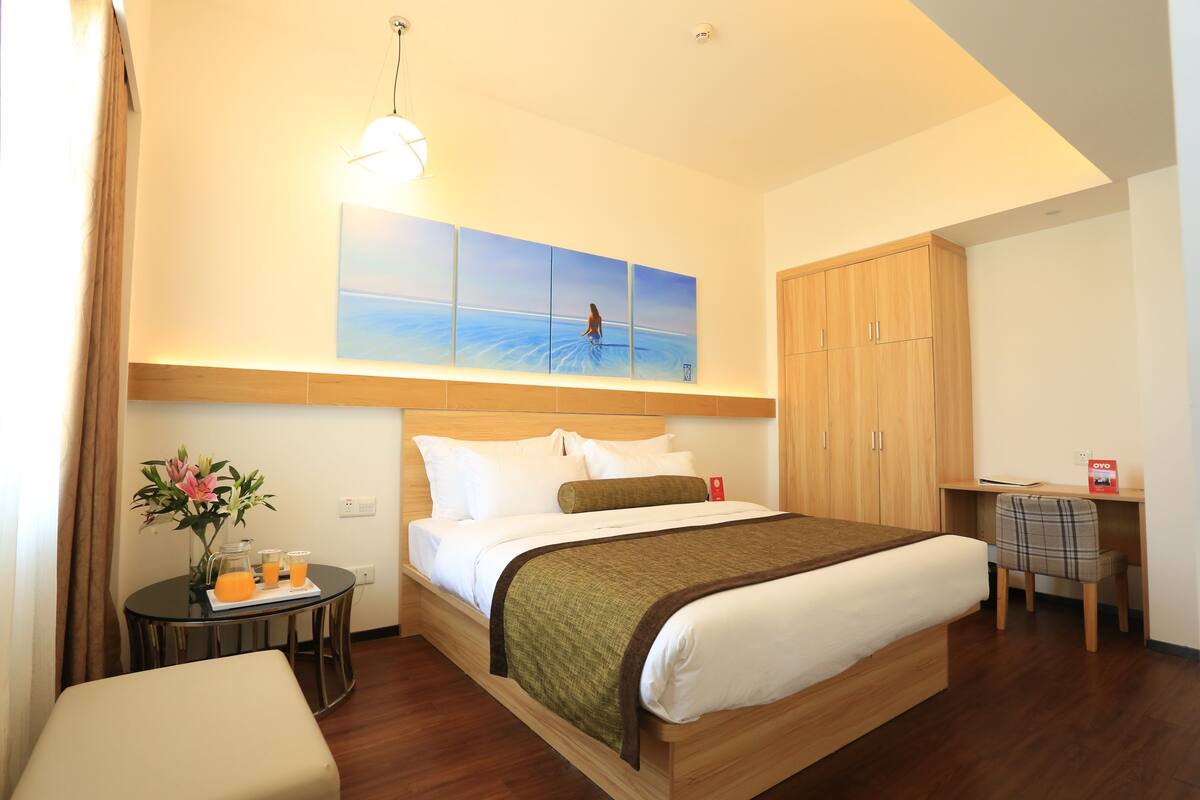 A hotel room with a bed, a desk, a chair, a small table with a vase and drinks, and a painting on the wall.