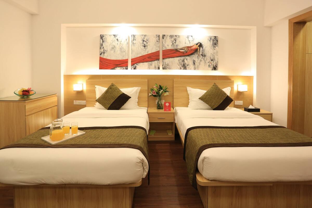 A hotel room with two beds, a wooden headboard, a painting above the beds, bedside lamps, a nightstand with a vase of flowers, and a tray with orange juice and glasses.