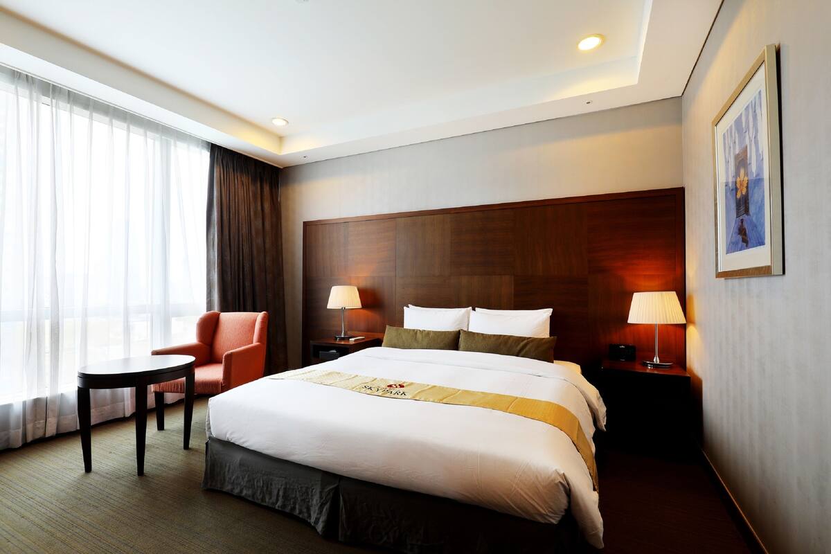 A hotel room with a large bed, a chair, a small table, and a nightstand with lamps.