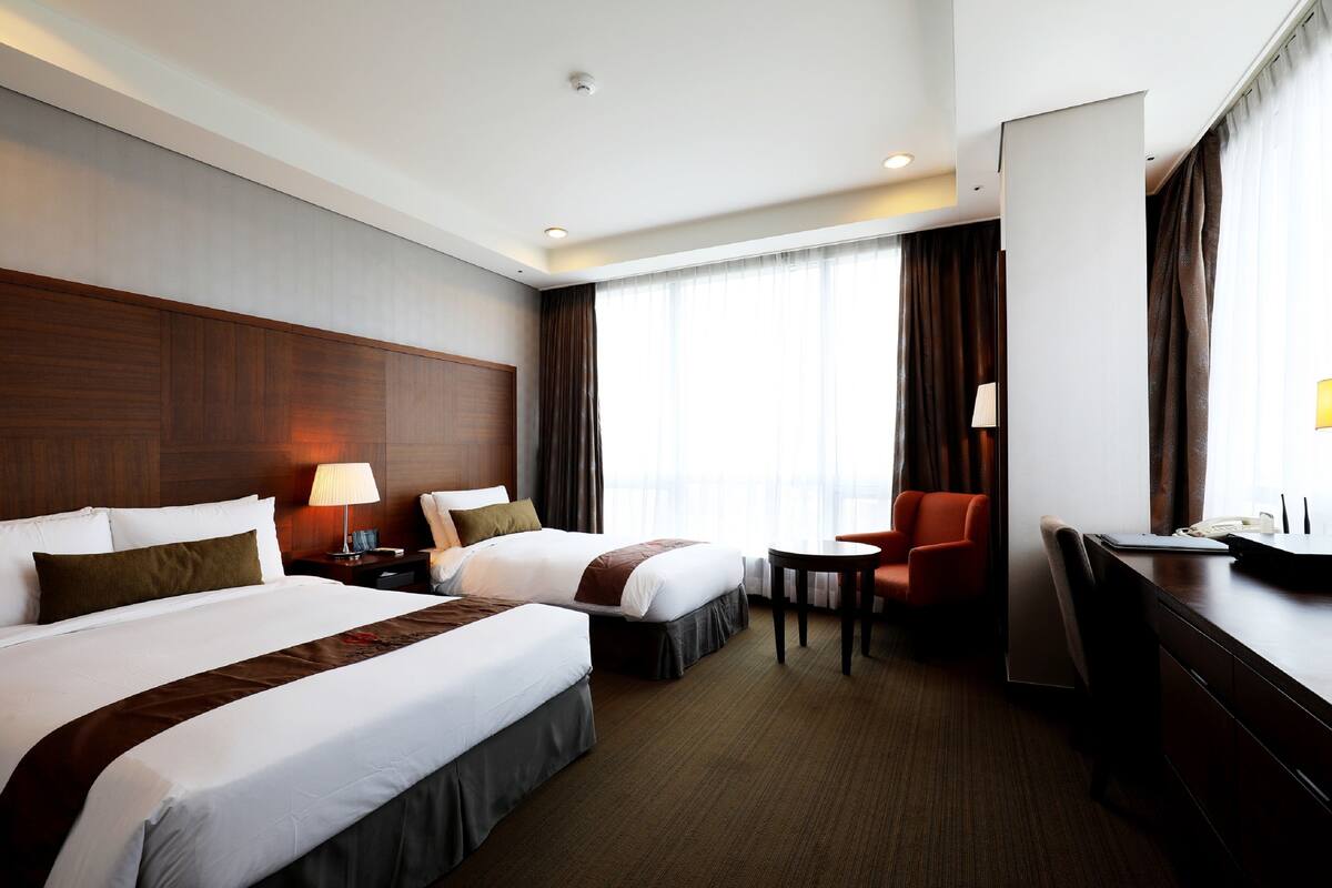 A hotel room with two beds, a desk, a chair, and a large window with curtains.