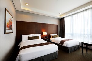 Habitación de hotel con dos camas, cabecero de madera, mesita de noche con lámpara y un ventanal con cortinas.