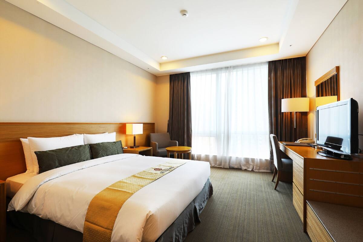 A hotel room with a large bed, a desk with a chair, a television, and a window with curtains.