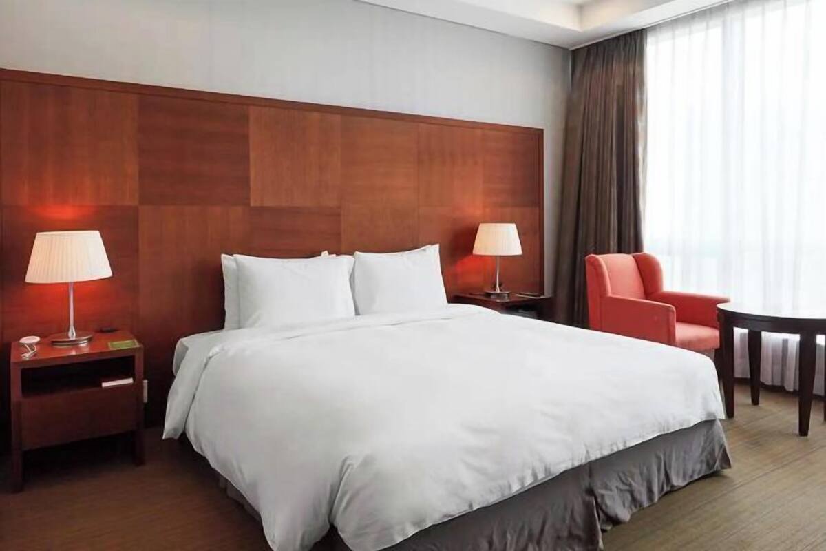 A hotel room with a large bed, two bedside lamps, a red chair, a small table, and a view of the outdoors through the window.
