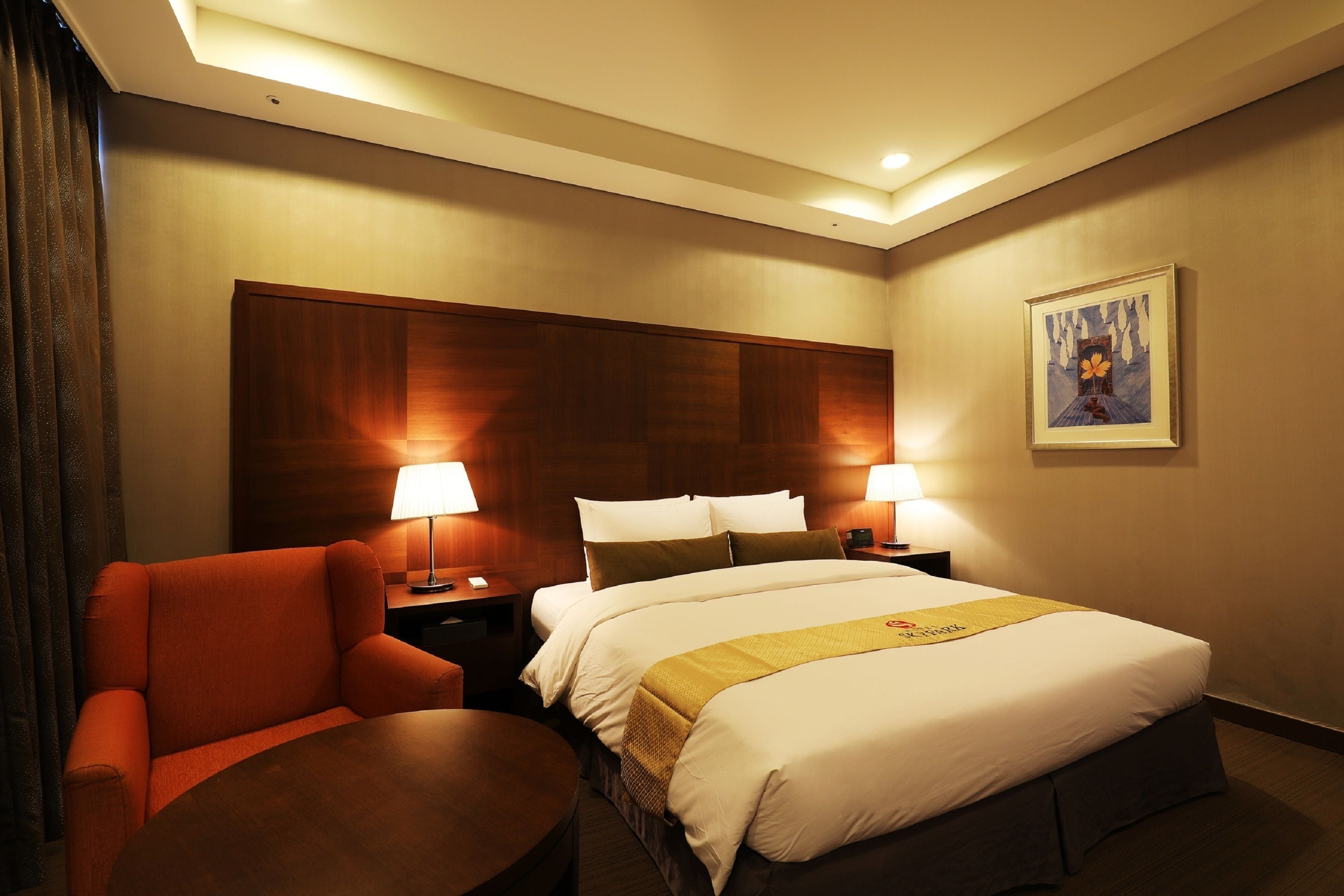 A hotel room with a large bed, two bedside lamps, a wooden headboard, a red armchair, a small round table, and a framed picture on the wall.