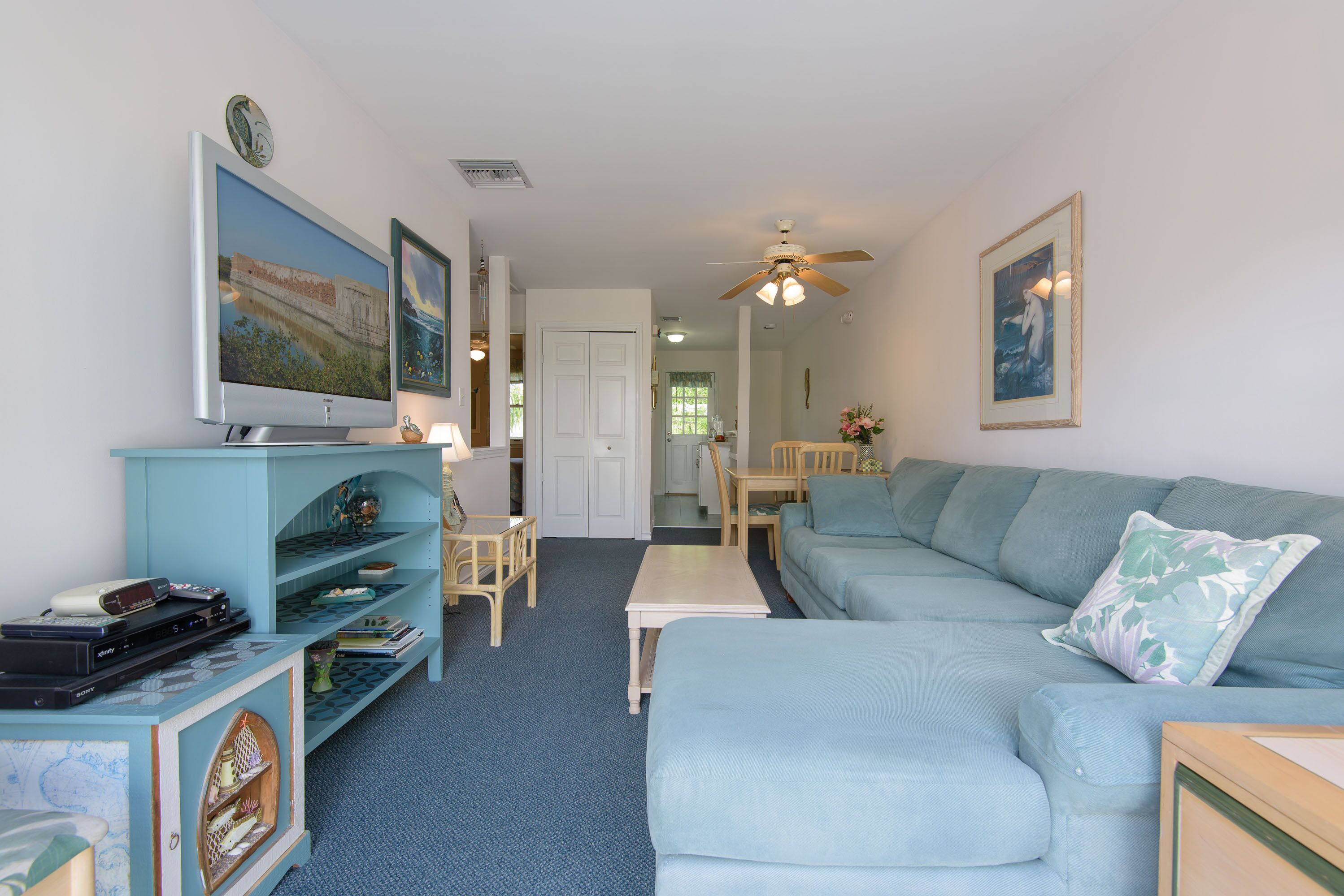 Ein Wohnzimmer mit einem blauen Sofa, einem Couchtisch und einem Fernseher auf einem Schrank. Ein Deckenventilator und eine Tür zu einem weiteren Raum sind ebenfalls vorhanden.