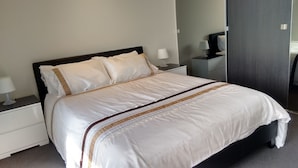 A neatly made bed with white linens and decorative trim, a bedside table with a lamp, and a mirror on the wall. - Camberwell Holiday Apartment (Camberwell)