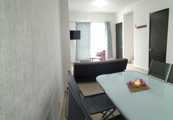 Une pièce moderne avec une table à manger en verre, un canapé, un téléviseur fixé au mur et une fenêtre avec des rideaux.