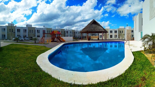 En swimmingpool med en paviljong med halmtak, omgiven av en välskött gräsmatta och moderna byggnader i bakgrunden.